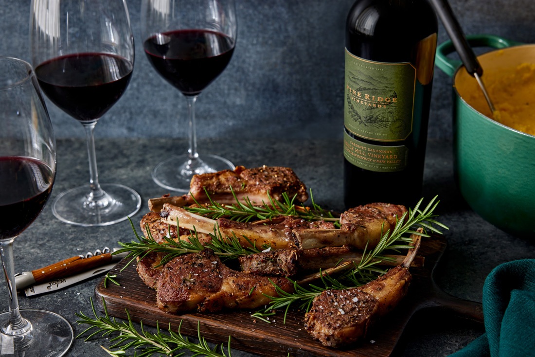 Grilled lamb chops garnished with rosemary are arranged on a wooden board. Three glasses of red wine and a bottle are nearby. A green pot is partially visible in the background on a dark surface.