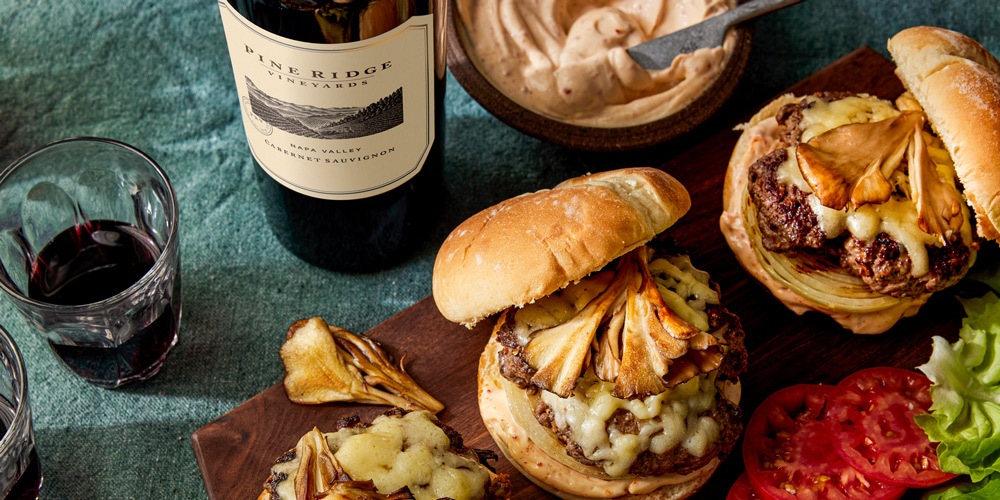 Two cheeseburgers with melted cheese and mushrooms on a wooden board, accompanied by sliced tomatoes, lettuce, and a bowl of sauce. A bottle of red wine labeled Penridge Vineyards and a glass of wine are in the background.