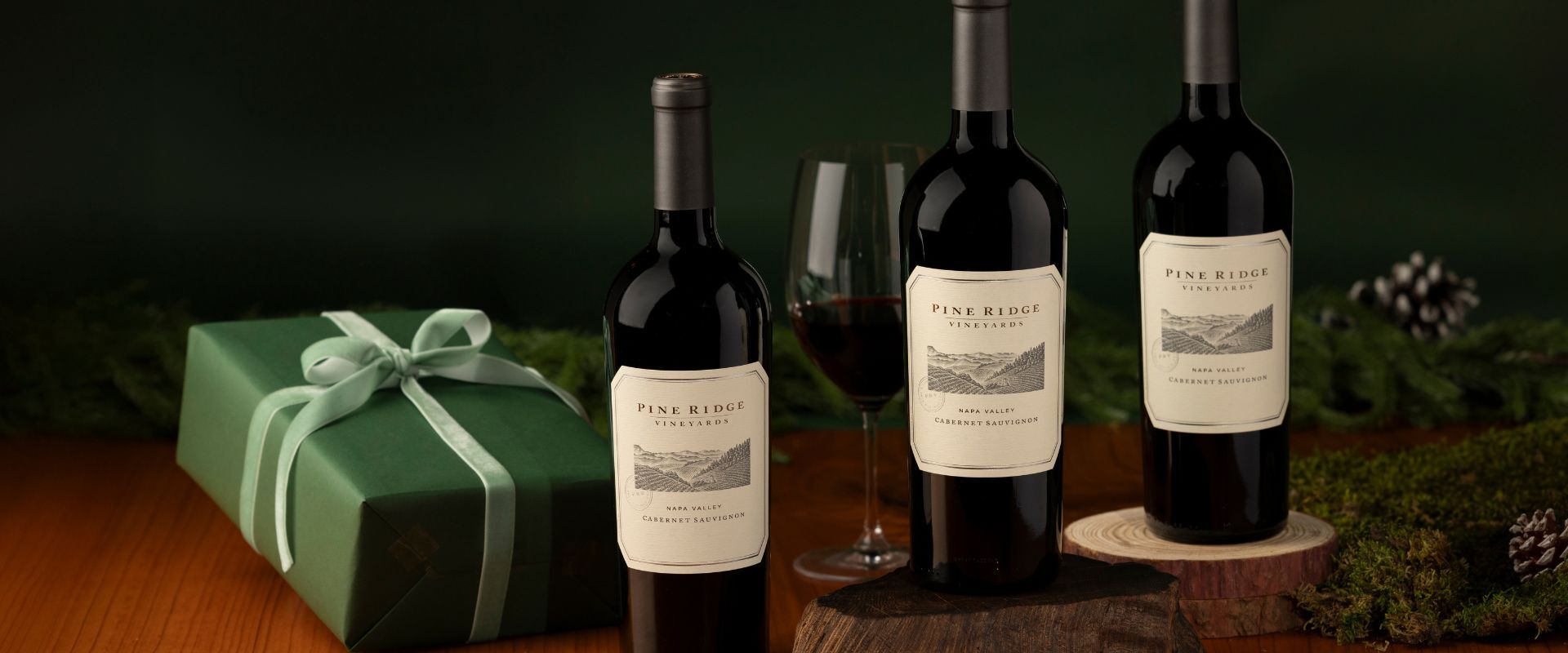 Three bottles of Pine Ridge Vineyards wine are displayed next to a filled wine glass and a green gift box with a ribbon, set on a wooden surface with greenery and pinecones in the background.