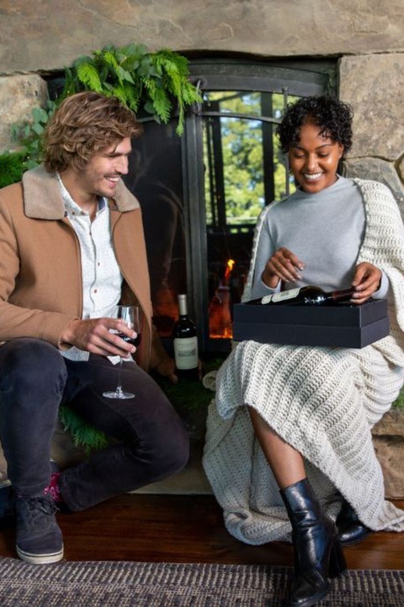 A man holding a glass of red wine sits beside a woman who is smiling and opening a black gift box. They are seated in front of a stone fireplace decorated with greenery, with a bottle of wine visible in the background.