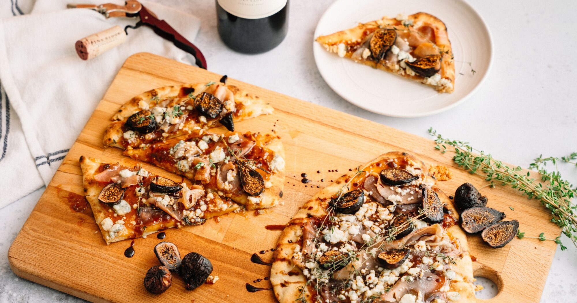 A wooden board holds sliced flatbread topped with figs, cheese, herbs, and drizzled sauce. Fresh figs and thyme are nearby. A small plate with a slice, a bottle of wine, cork, and towel are in the background.
