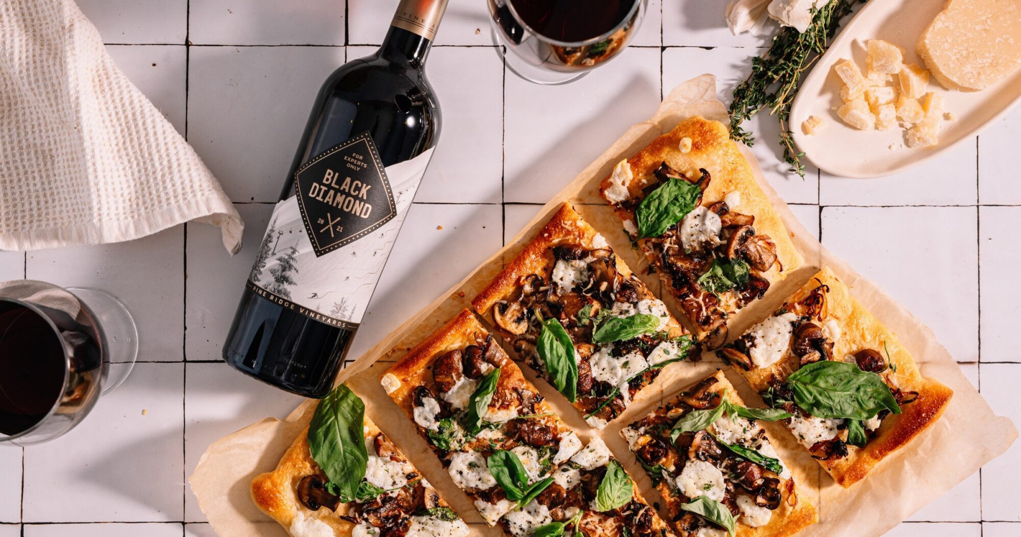 Rectangular pizza topped with cheese, mushrooms, olives, and basil, sliced into squares on parchment paper. A bottle of Black Diamond wine, two glasses of red wine, and a plate with cheese and herbs are nearby on a tiled surface.
