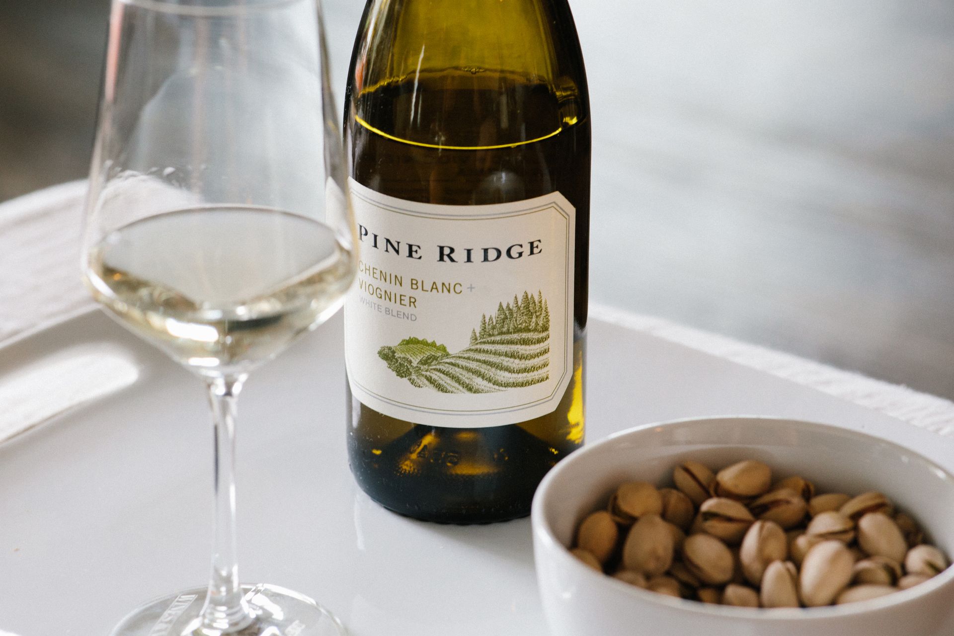 A glass and bottle of Pine Ridge Chenin Blanc + Viognier white wine are on a white surface next to a small white bowl filled with pistachios.