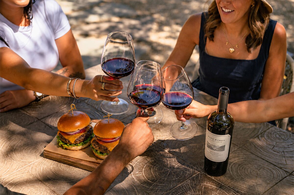 Four people toast with glasses of red wine around a wooden table with two burgers on a board and a bottle of wine. The group appears to be enjoying an outdoor meal on a sunny day.