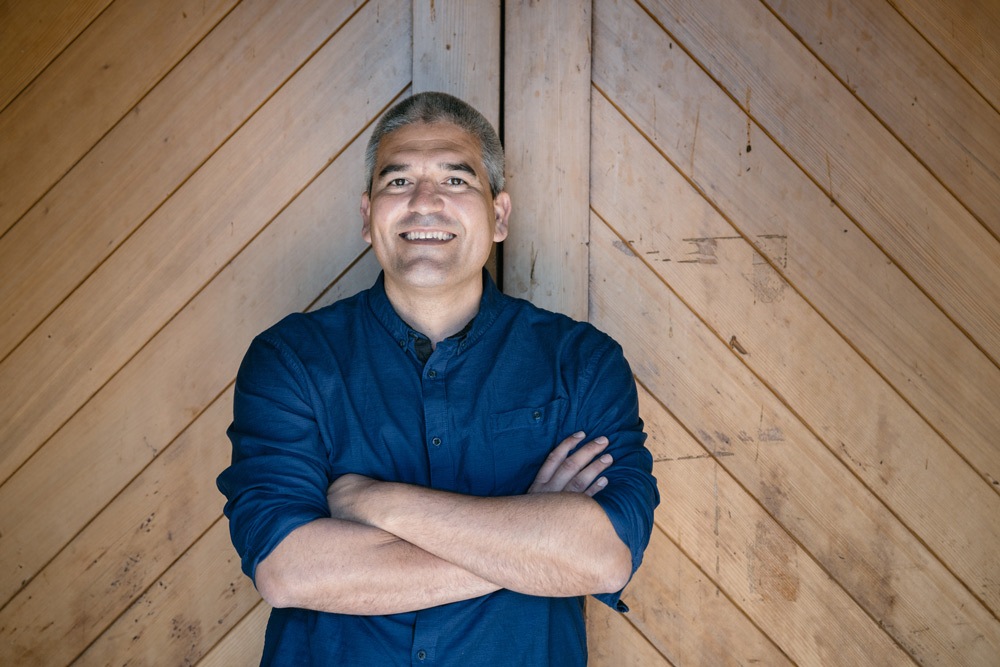 Josh Widaman is standing with arms crossed in front of wooden paneling. The person is smiling and looking at the camera. The background consists of diagonal wooden planks.
