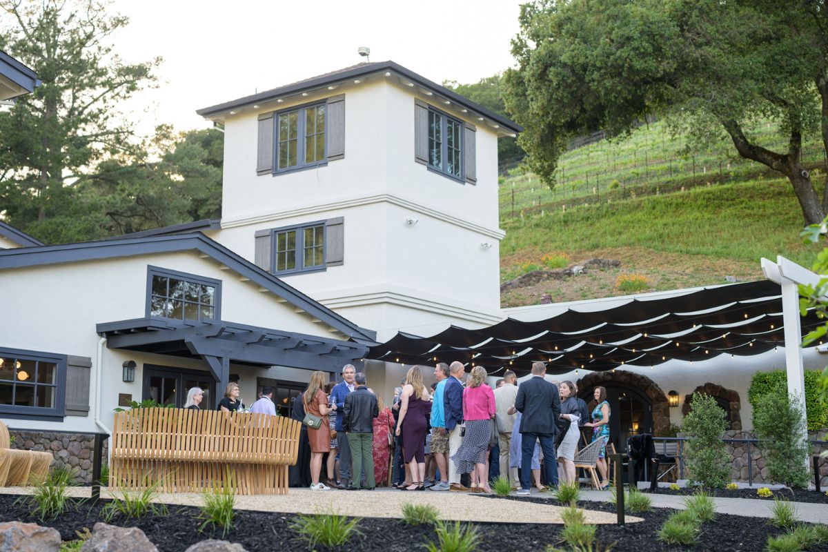 A group of people gather outdoors at an event near a large house with a modern design. They are standing on a patio with a canopy, surrounded by greenery and trees. A bar area is visible to the left.