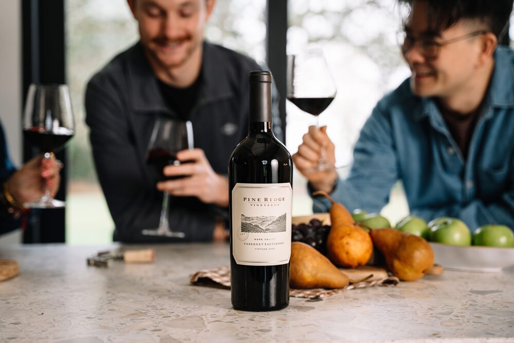 A bottle of Pine Ridge Vineyards Cabernet Sauvignon is on a countertop, surrounded by pears, grapes, and apples. Two people in the background hold glasses of red wine and appear to be talking.
