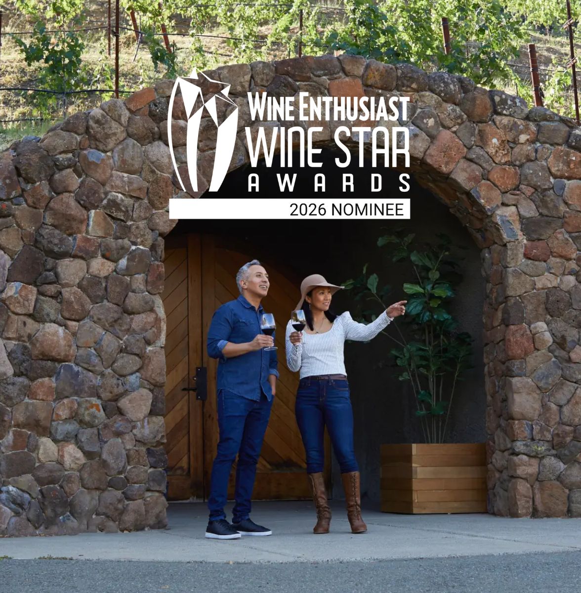 Two people stand outside a stone wine cellar holding glasses of wine, with the text Wine Enthusiast Wine Star Awards 2026 Nominee displayed above them. Both are casually dressed and facing right.