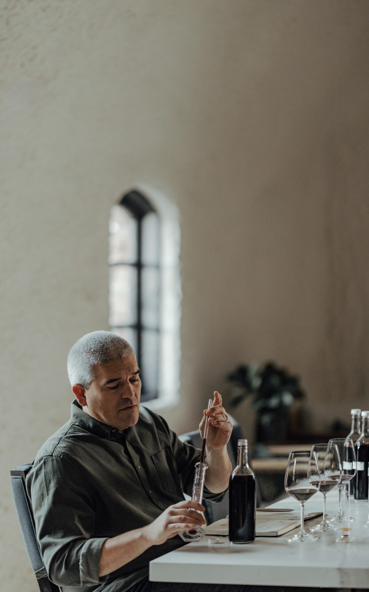 A man with short gray hair sits at a table, using a dropper to add liquid to a glass container. There are bottles, wine glasses, and a window with arched framing in the background.