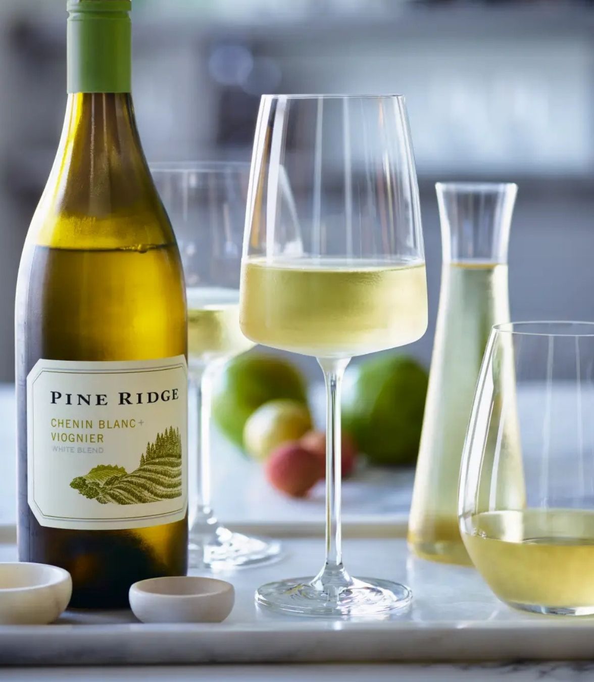A bottle of Pine Ridge Chenin Blanc + Viognier wine is on a marble surface. Two wine glasses, one filled and one partially filled, are next to it. In the background, there are blurred fruits and a carafe with liquid.