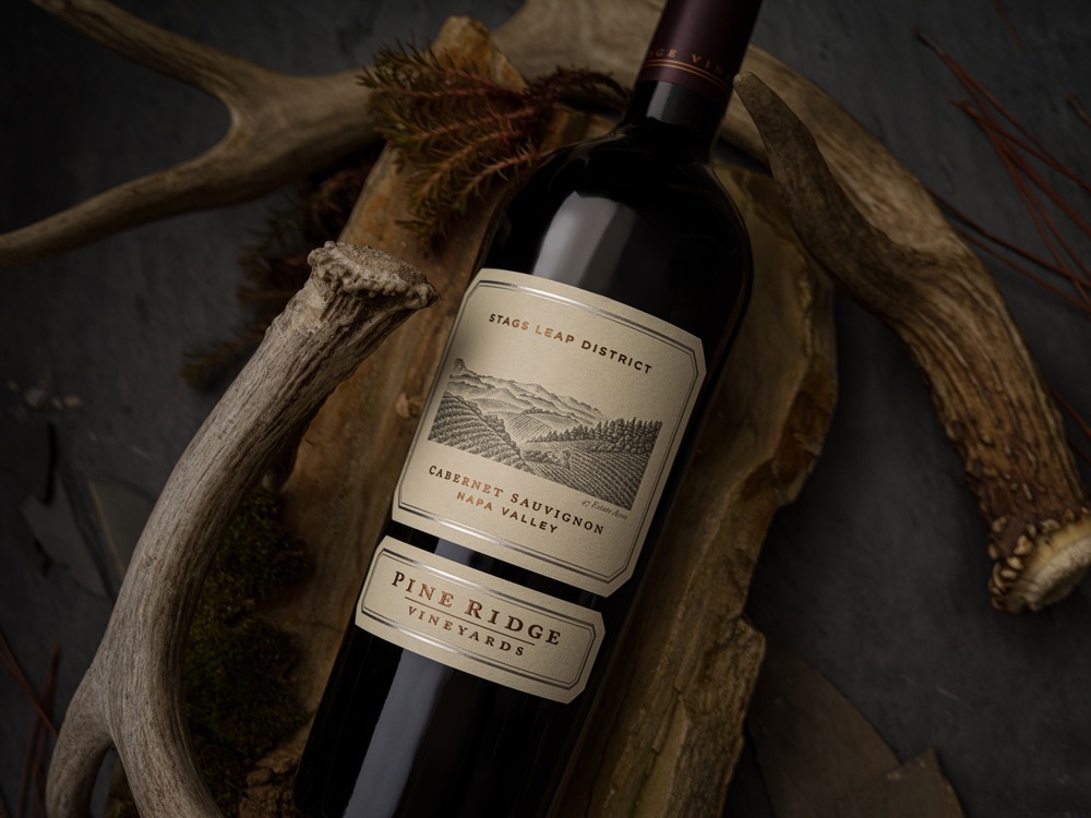 A bottle of Pine Ridge Vineyards Cabernet Sauvignon from the Stags Leap District of Napa Valley rests on a wooden surface with antlers. The label features a landscape illustration. Pine needles are scattered around.