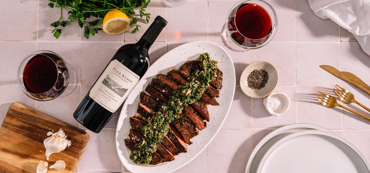A platter of sliced steak topped with chimichurri sauce sits on a table. Beside it are two glasses of red wine, a wine bottle, garlic, a lemon, herbs, and a small bowl of pepper. Plates and gold-colored cutlery are on the right.