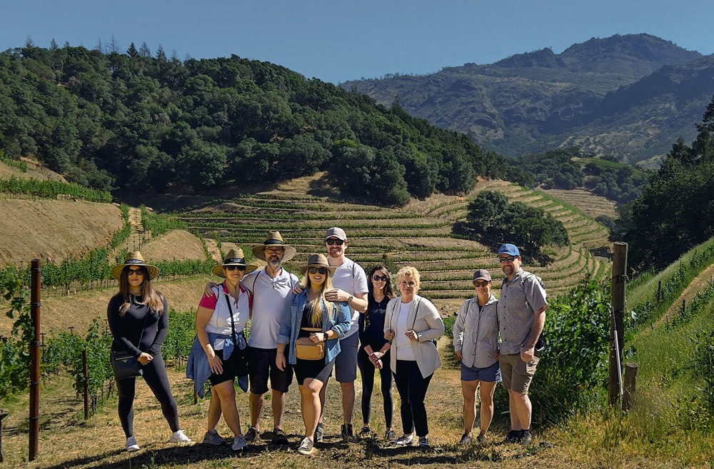 A group of nine people stand outdoors in front of a vineyard on a sunny day. They are wearing casual clothing and hats, with a backdrop of terraced grapevines and rolling hills.