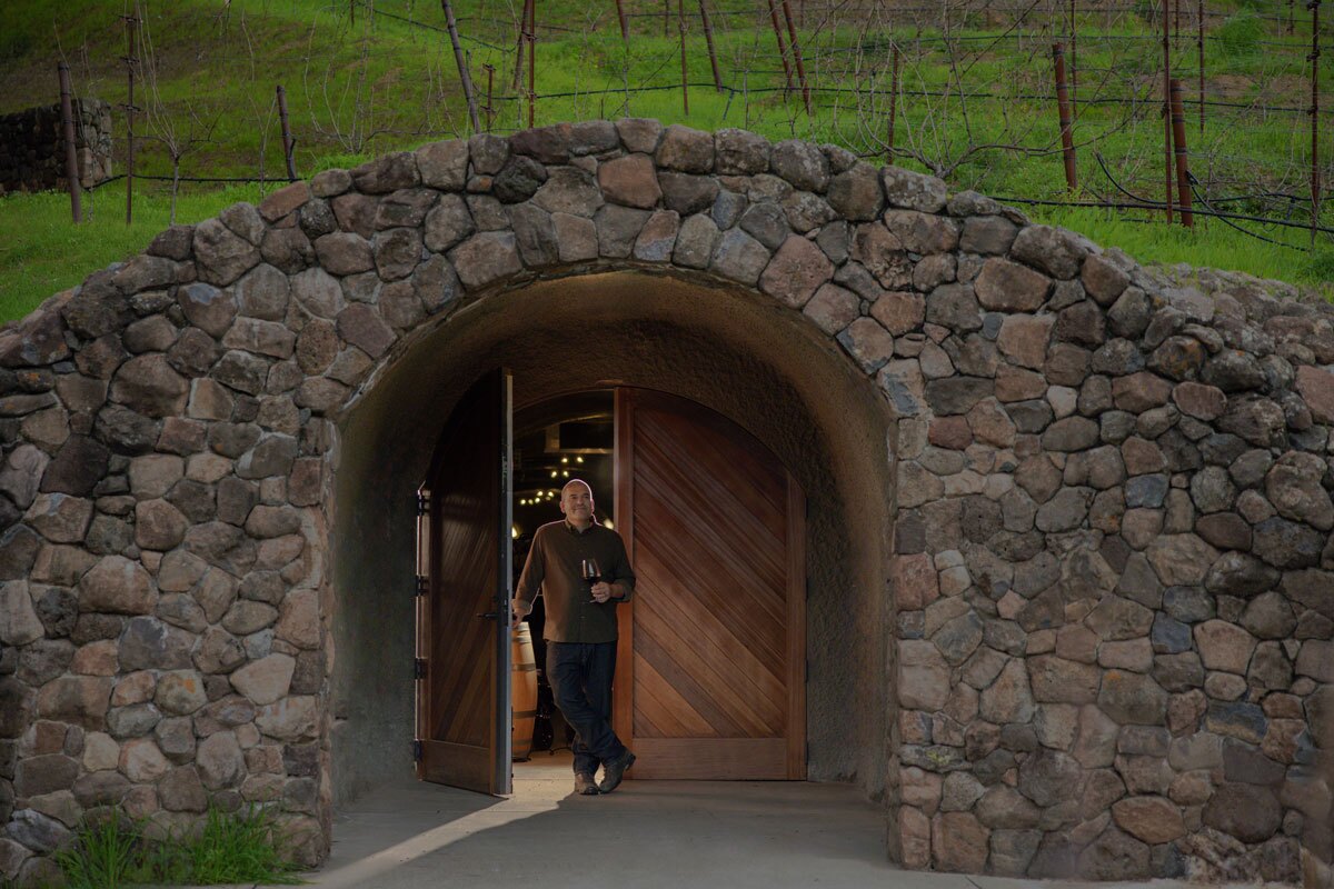 Winemaker, Josh Widaman, stands at the entrance of a stone cellar, holding a wine glass. The arched doorway is made of wood. The background shows a lush green hillside with a vineyard. The scene is serene and indicative of a vineyard or winery setting.