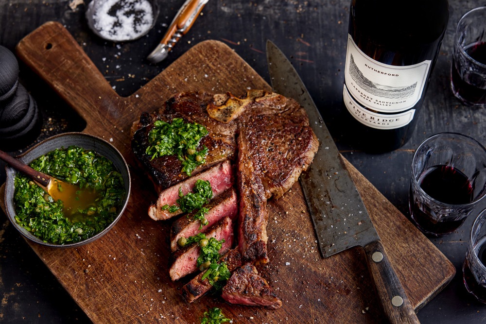 A sliced cooked steak with green sauce on top sits on a wooden cutting board next to a large knife. A bowl of sauce, a bottle of red wine, two glasses, and salt are nearby.