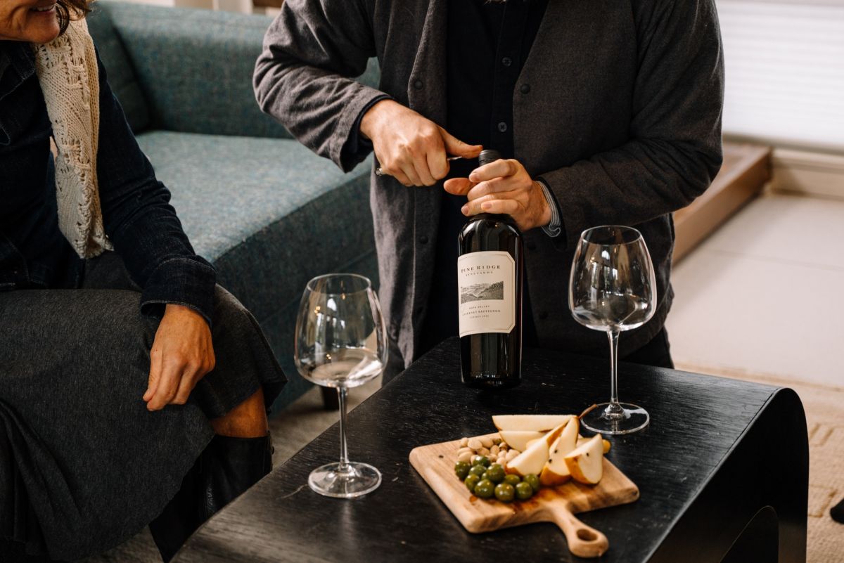 A person opens a bottle of wine with a corkscrew at a table set with two empty wine glasses, a wooden board with cheese, olives, and crackers, while another person sits nearby.