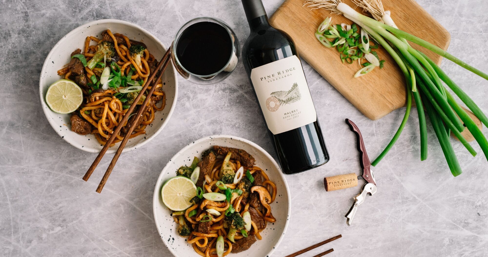 Two bowls of stir-fried noodles with vegetables and lime slices, a bottle of red wine with a filled glass, a cork and corkscrew, and a cutting board with sliced green onions on a gray surface.