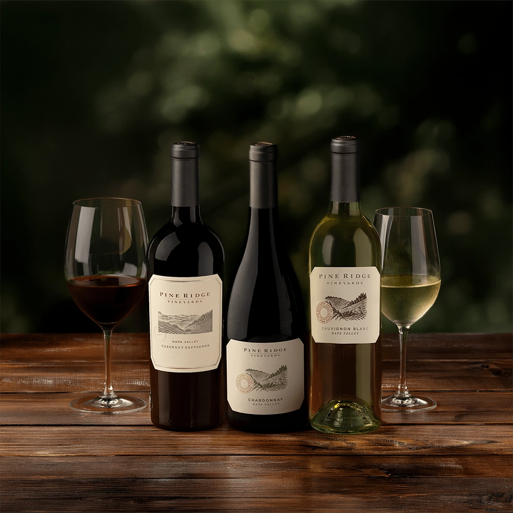 The Curated Trio: Three bottles of Pine Ridge wine—one red, one white, and one Chardonnay—stand on a wooden table, each beside a filled glass, with a green, blurred background.