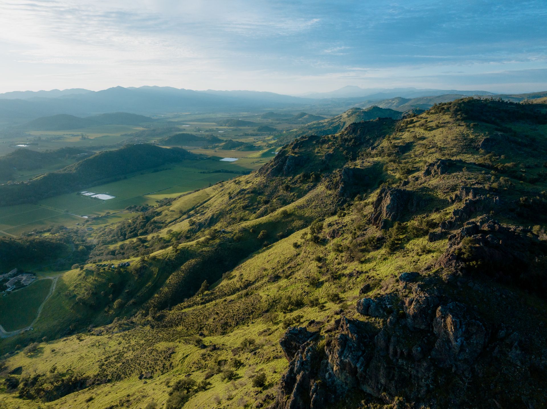 Aerial view of a green, hilly landscape with rocky ridges, patches of farmland, and distant mountains under a partly cloudy sky.