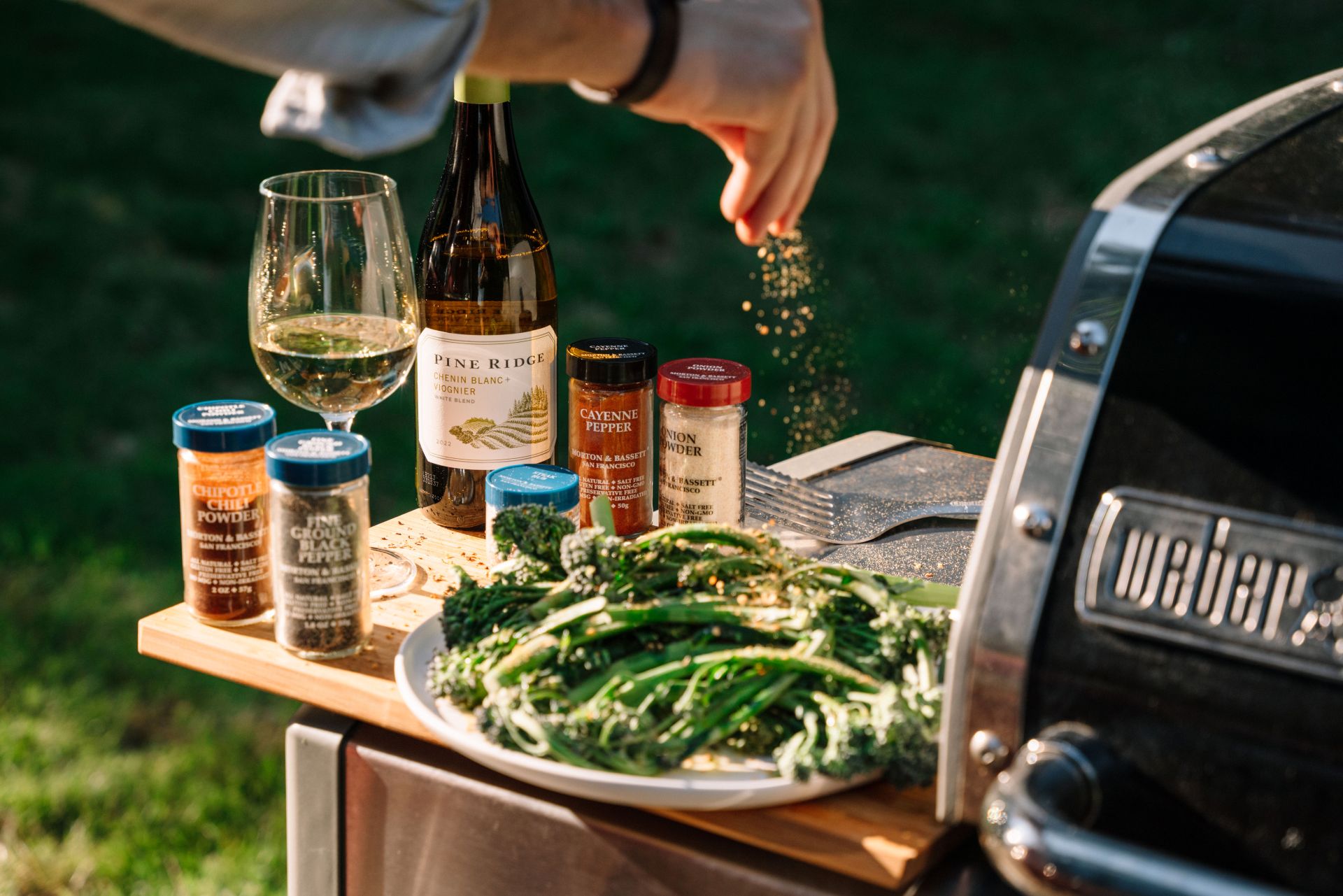 A person sprinkles seasoning onto a plate of broccolini on an outdoor grill station, which also holds bottles of wine, a glass of white wine, spice containers, and grilling utensils.