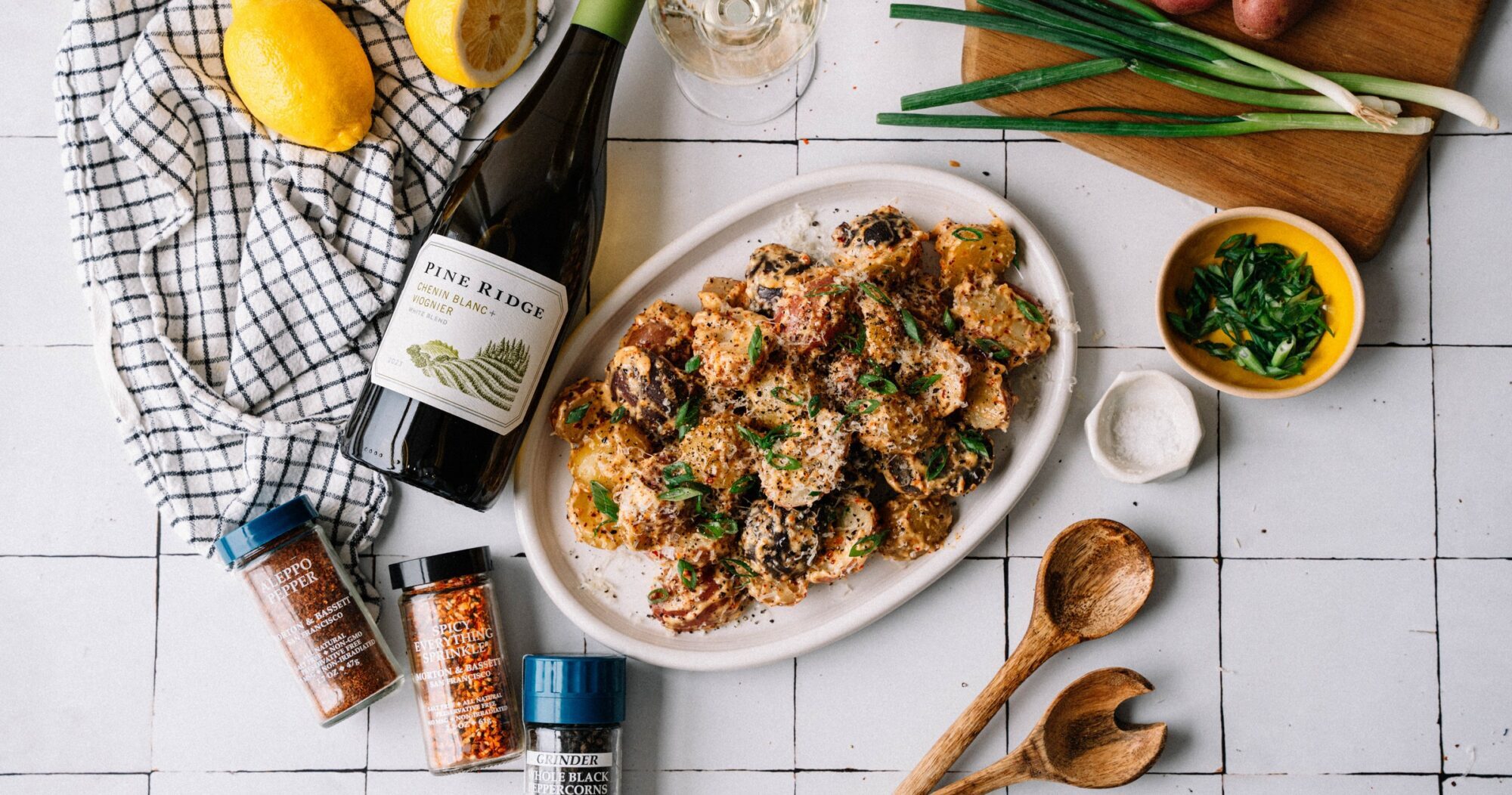 A platter of roasted potatoes garnished with herbs sits on a white table next to a bottle of wine, wooden utensils, spices, green onions, lemons, and a checkered cloth.