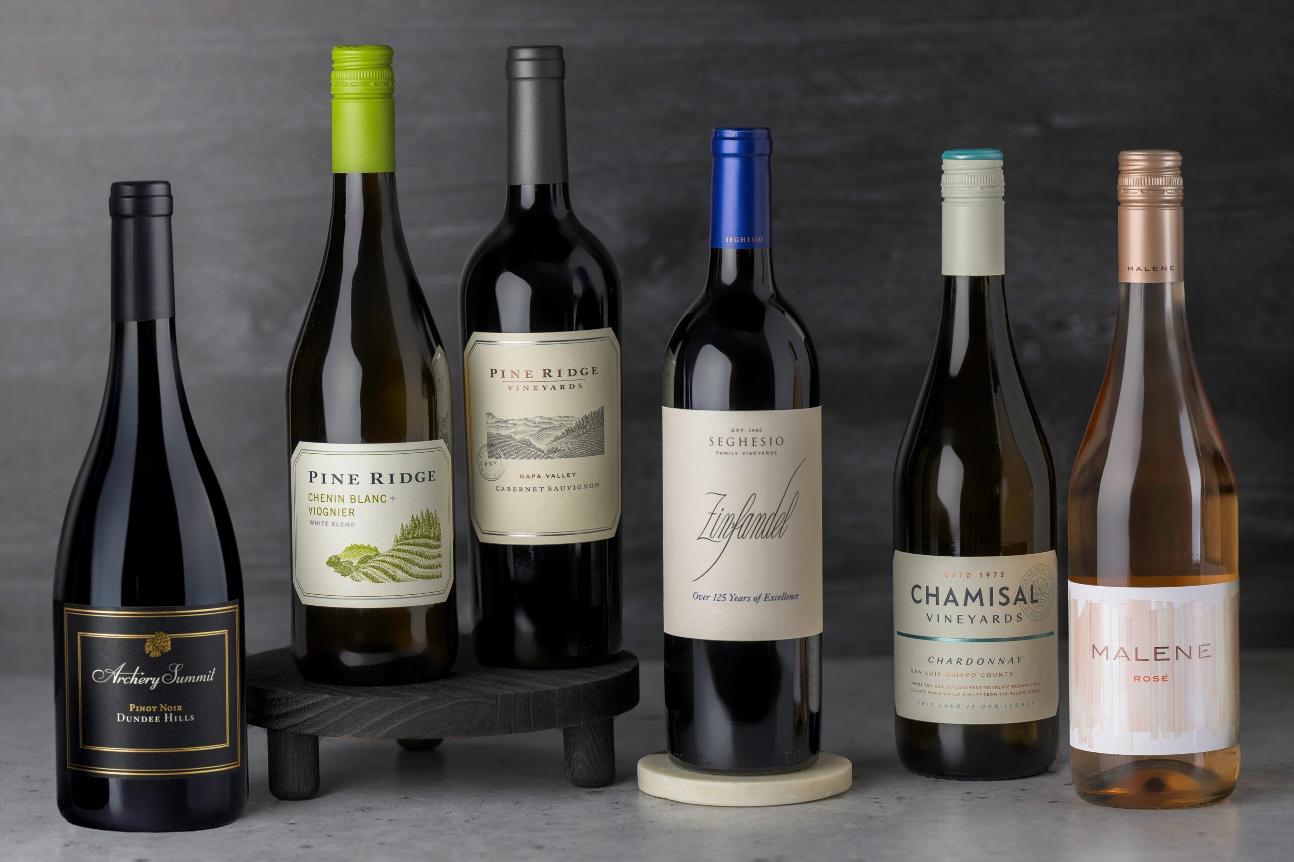Six bottles of wine, each with distinct labels, are arranged upright on a surface against a gray background. One bottle sits on a small black platform, and the wines include red, white, and rosé varieties.