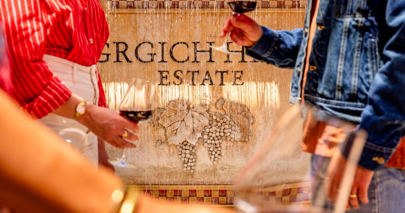 Three people holding glasses of red wine stand in front of a stone fountain with the engraved words GRGICH HILLS ESTATE and a grapevine motif. Only their torsos and arms are visible.