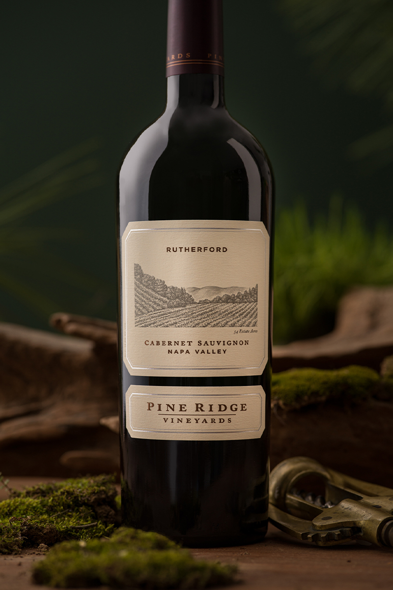 A bottle of Pine Ridge Vineyards Cabernet Sauvignon from Napa Valley is placed upright. The label has an illustration and the word Rutherford on it. Moss and a vintage corkscrew surround the bottle, set against a green background.