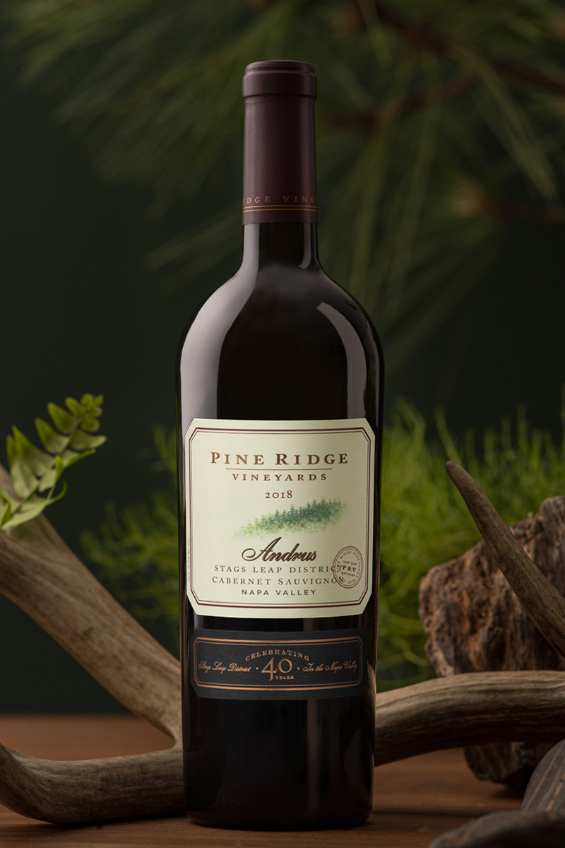 A bottle of Pine Ridge Vineyards wine is displayed. The label reads 2018 Stags Leap District Cabernet Sauvignon, Napa Valley. The background includes green foliage and a wooden element.