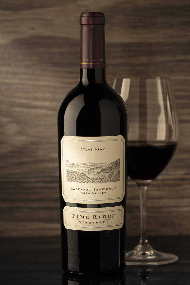 A bottle of Pine Ridge Vineyards Cabernet Sauvignon, labeled Atlas Peak and Napa Valley, is placed next to a filled wine glass on a dark surface with a wooden background.