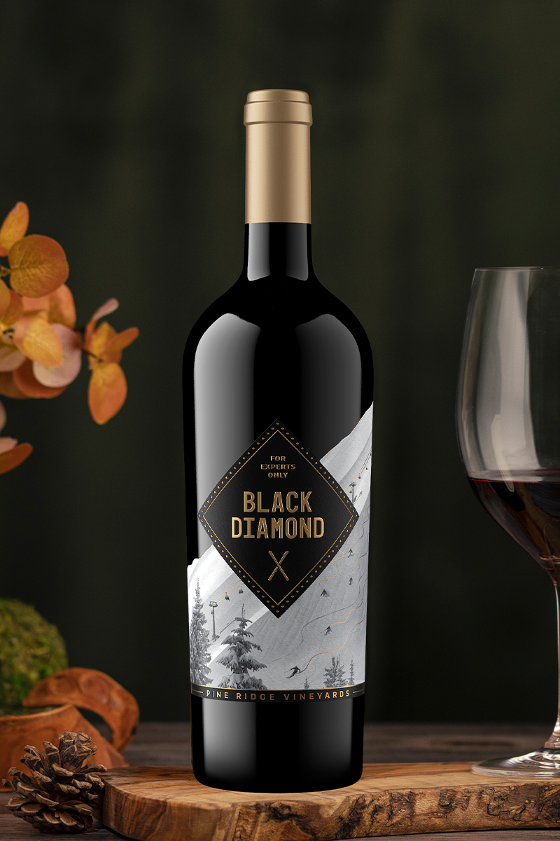 A bottle of Black Diamond wine is displayed on a wooden surface next to a glass of red wine. The label features a silver mountain landscape design. Autumn leaves and a small green ornament are blurred in the background.