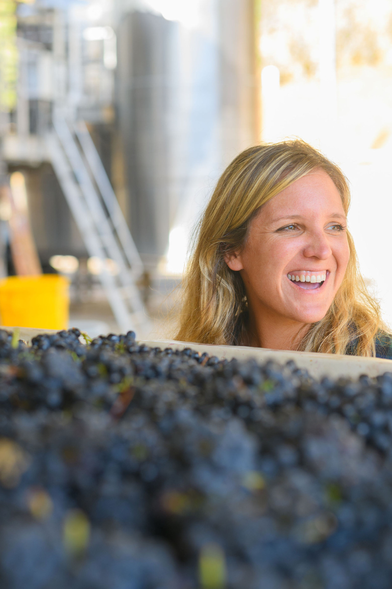 A woman with long blonde hair smiles while standing behind a large container filled with dark grapes at an industrial facility.