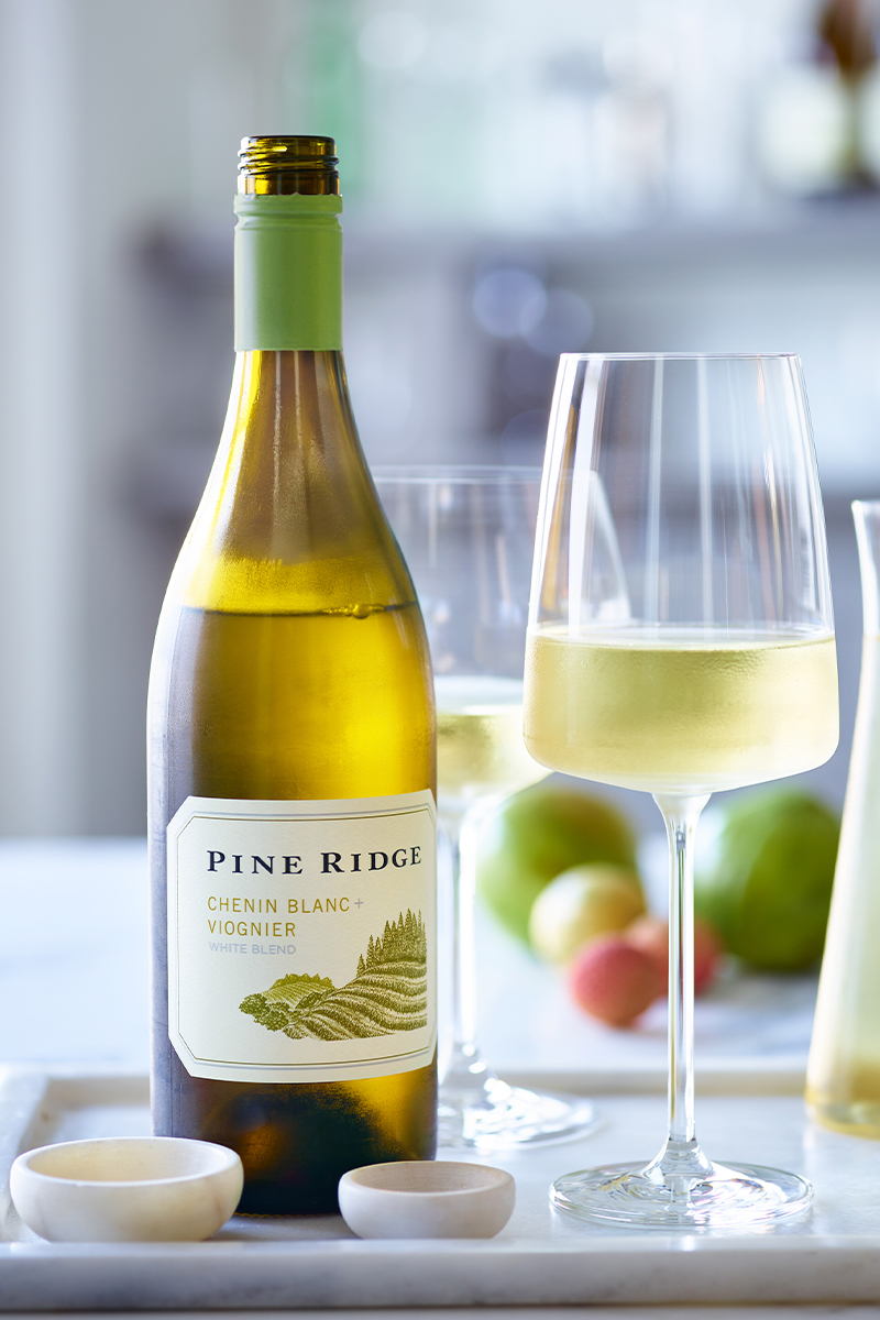 A bottle of Pine Ridge Chenin Blanc + Viognier wine is placed on a table next to a filled wine glass. The table has blurred fruits in the background and two small bowls in the foreground.