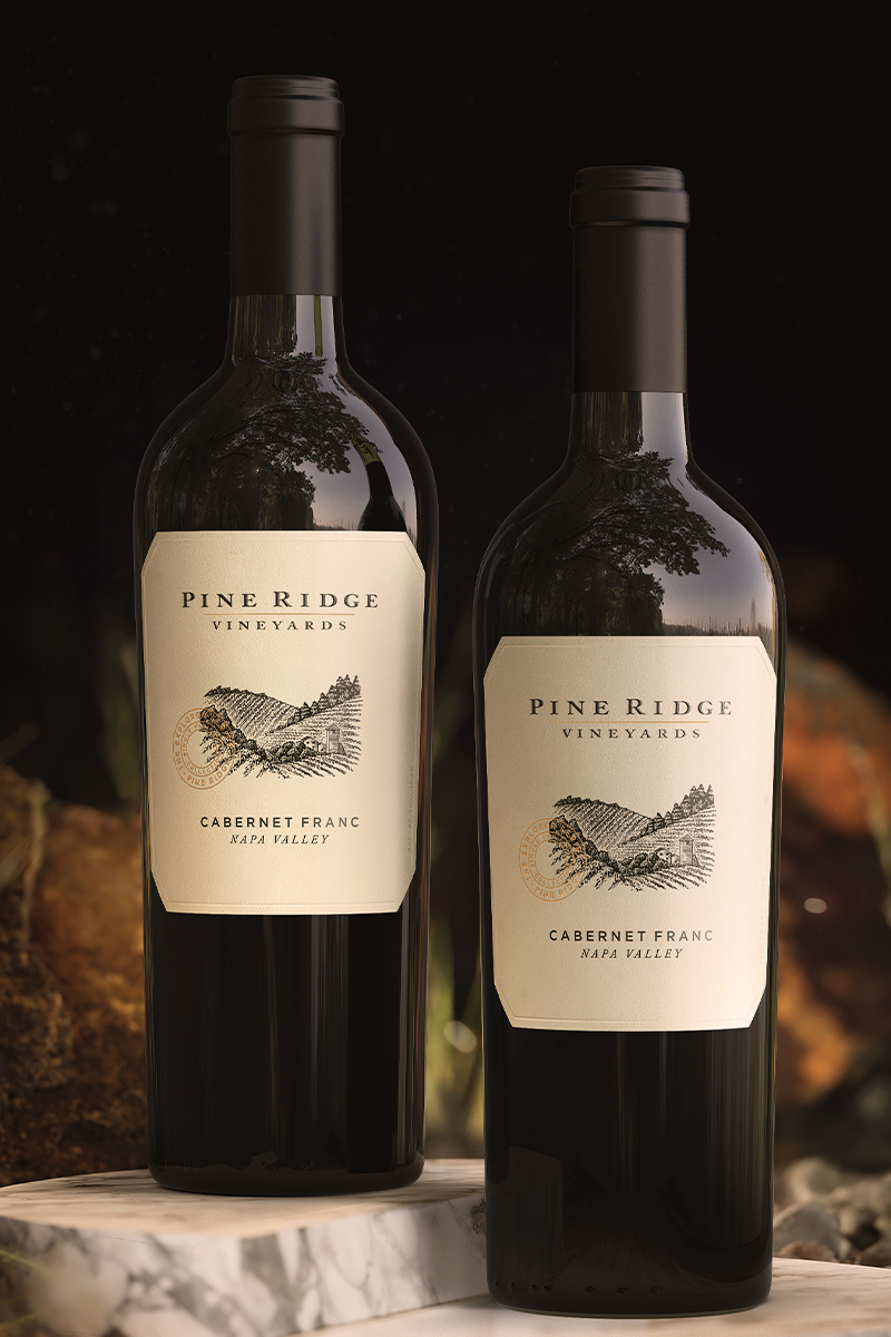 Two bottles of Pine Ridge Vineyards Cabernet Franc wine stand side by side on a marble surface. Each label showcases detailed winery information and an illustrated pine ridge landscape, perfectly complementing the soft-focus natural setting in the background.