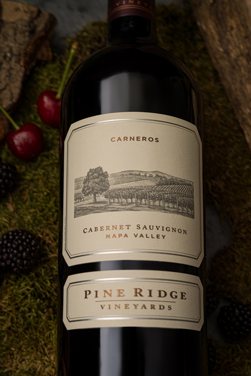A bottle of Pine Ridge Vineyards Cabernet Sauvignon from Napa Valley is placed on moss with cherries and blackberries nearby. The label features an illustration of a vineyard landscape and the word Carneros.