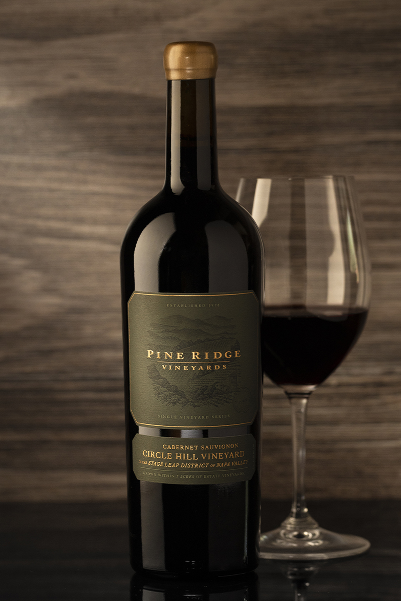 A bottle of Pine Ridge Vineyards Cabernet Sauvignon stands beside a filled wine glass. The label includes details about the vineyard and its age. The background has a textured, wooden appearance.