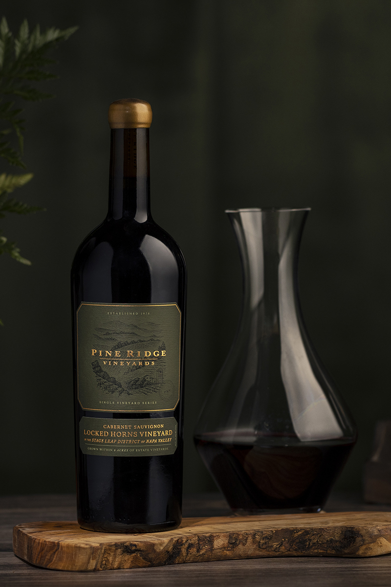 A wine bottle with a label reading Pine Ridge Vineyards and Locked Horns Cabernet Sauvignon stands next to a filled glass decanter on a wooden surface. The background is a dark, blurred green, with partial plants visible on the side.