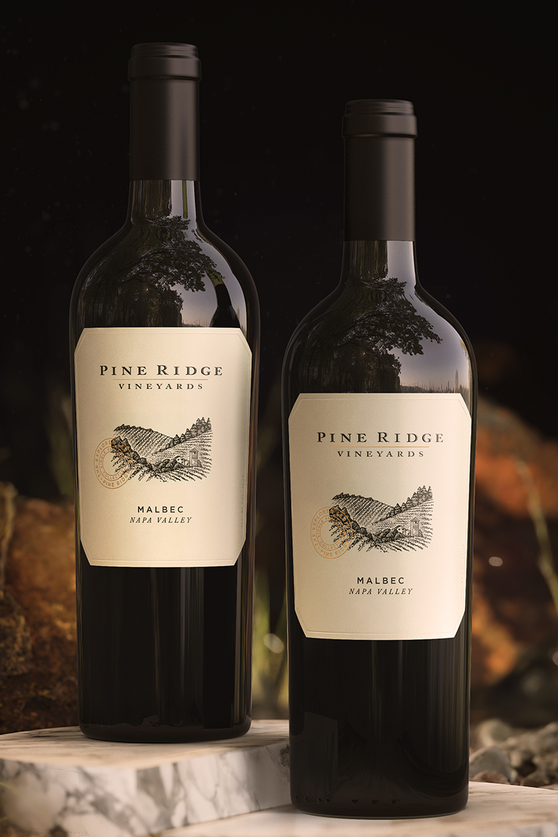 Two bottles of Malbec from Pine Ridge Vineyards in Napa Valley rest on a marble surface. Each bottle features a cream-colored label with an illustration of a ridge and the vineyards name. A softly blurred background completes this elegant display.