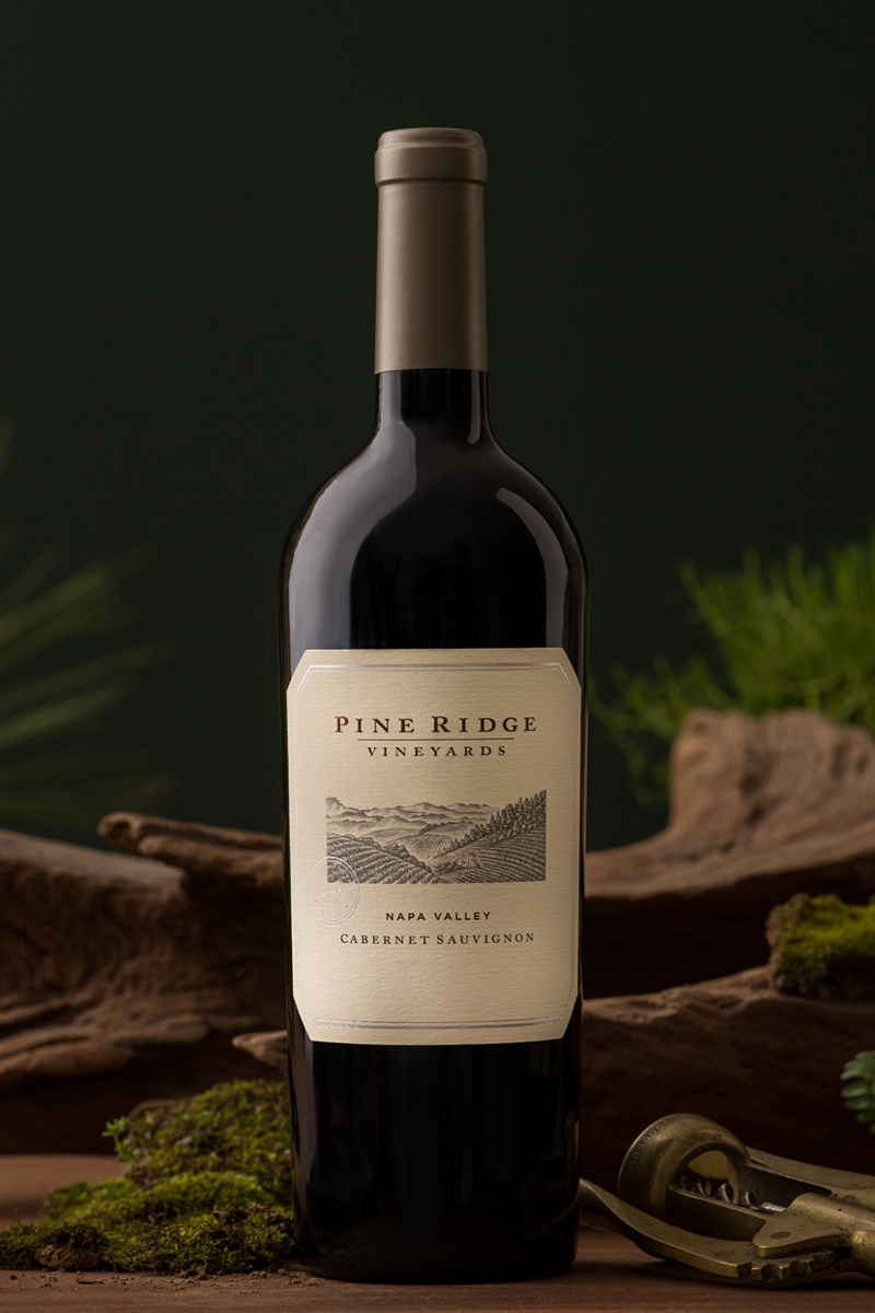 A bottle of Pine Ridge Vineyards Cabernet Sauvignon from Napa Valley stands on a wooden surface. The background includes green foliage and a dark green wall, with a rustic tool nearby.
