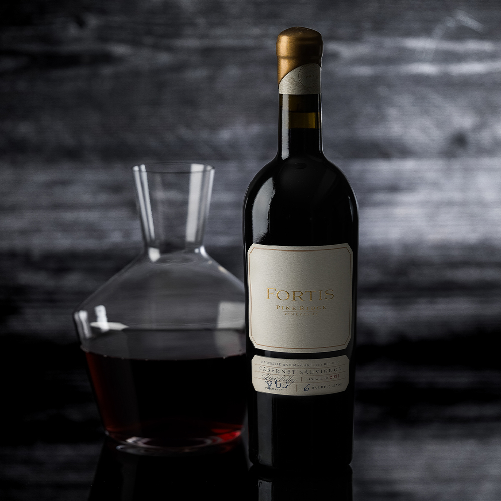 A bottle of Fortis Cabernet Sauvignon wine stands next to a glass decanter filled with red wine, set against a dark, textured background.