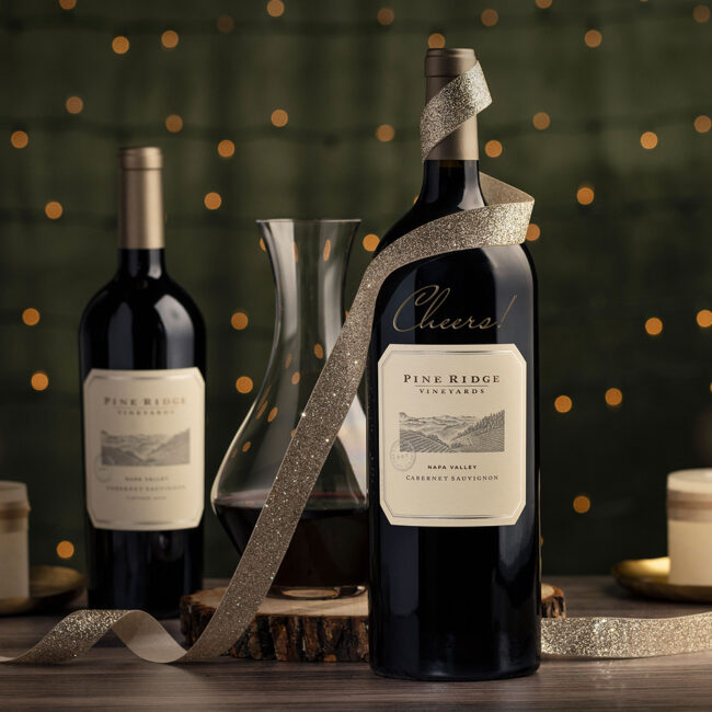 Two bottles of Pine Ridge Vineyards wine, a decanter, a wood slice, and candles are arranged on a table. One bottle is wrapped with a gold ribbon that says Cheers! Small lights are blurred in the green background.