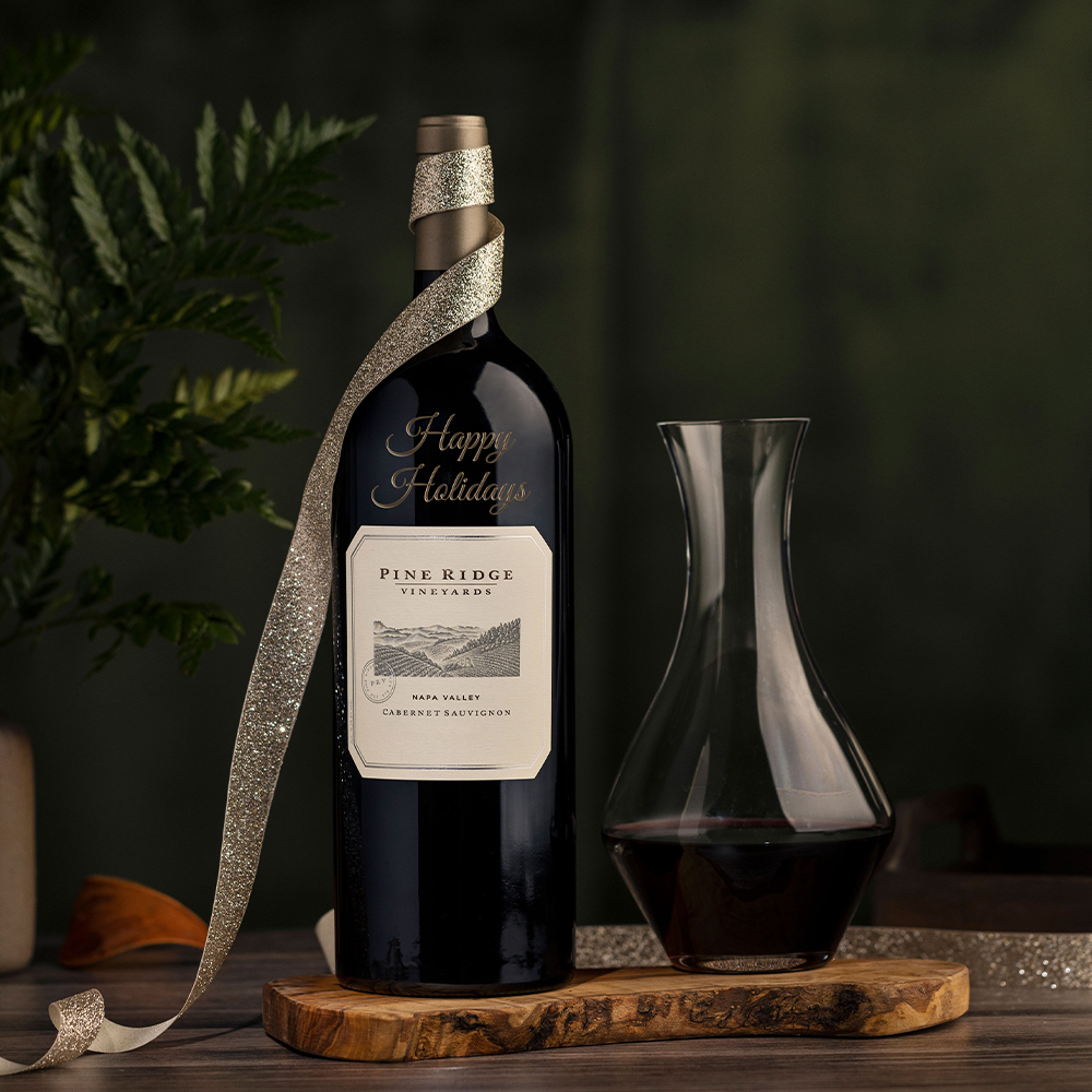 A bottle of Pine Ridge Vineyards Cabernet Sauvignon with a “Happy Holidays” message, wrapped in a silver ribbon, sits next to a glass decanter filled with wine on a wooden board against a dark, festive background.