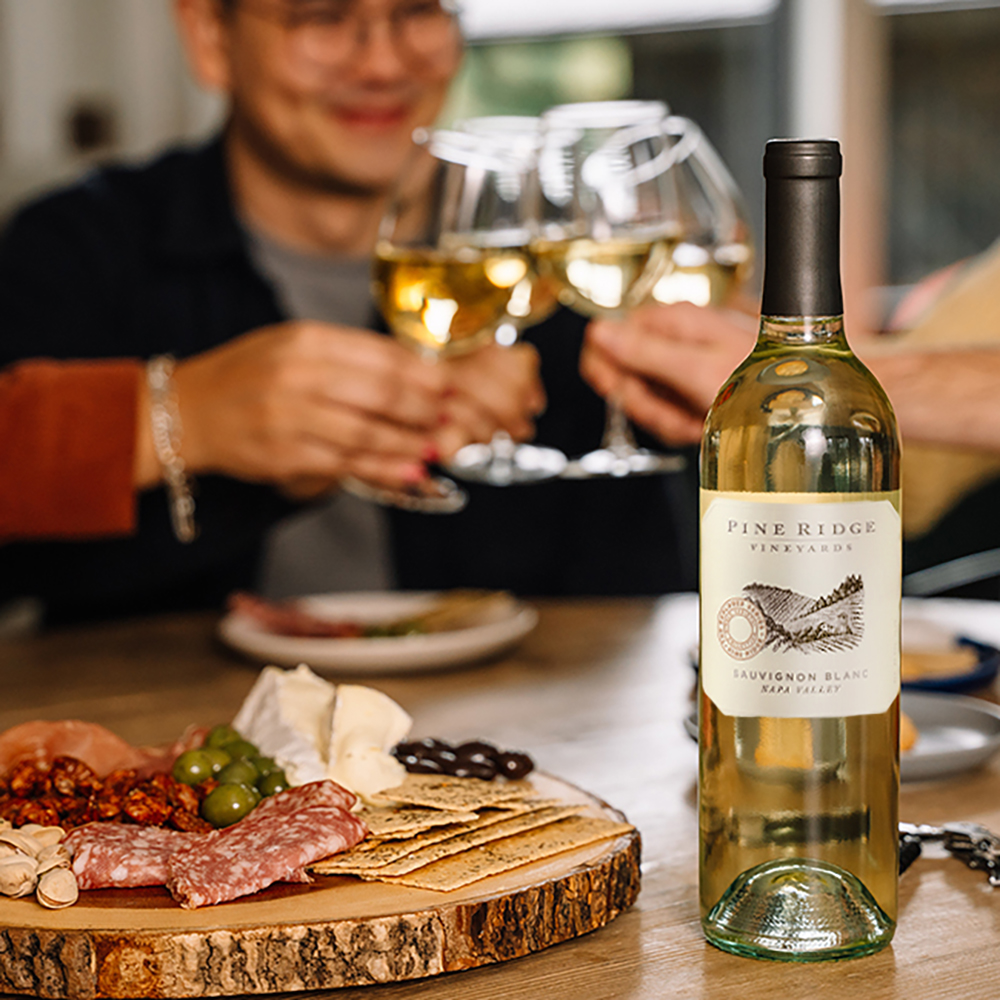 A bottle of Pine Ridge Vineyards Sauvignon Blanc sits on a table beside a charcuterie board with meats, cheeses, crackers, and olives, while friends in the background clink glasses of crisp Sauvignon Blanc.
