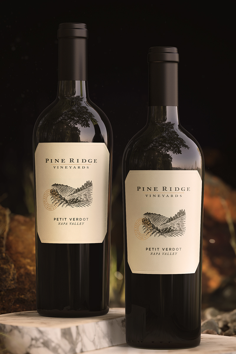 Two bottles of Pine Ridge Vineyards Petit Verdot wine stand upright on a white marble surface. Their beige labels, adorned with black text and a sketch of a vineyard landscape, catch the eye. The background is dark, enriched with subtle foliage, enhancing the elegance of these Petit Verdot wines.
