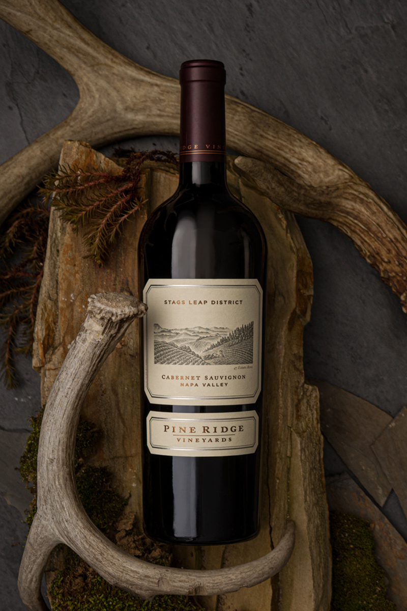 A bottle of Pine Ridge Vineyards Cabernet Sauvignon from Stags Leap District, Napa Valley, is placed on a backdrop of antlers and wood. The bottle is upright, with a label depicting a vineyard scene in monochrome.