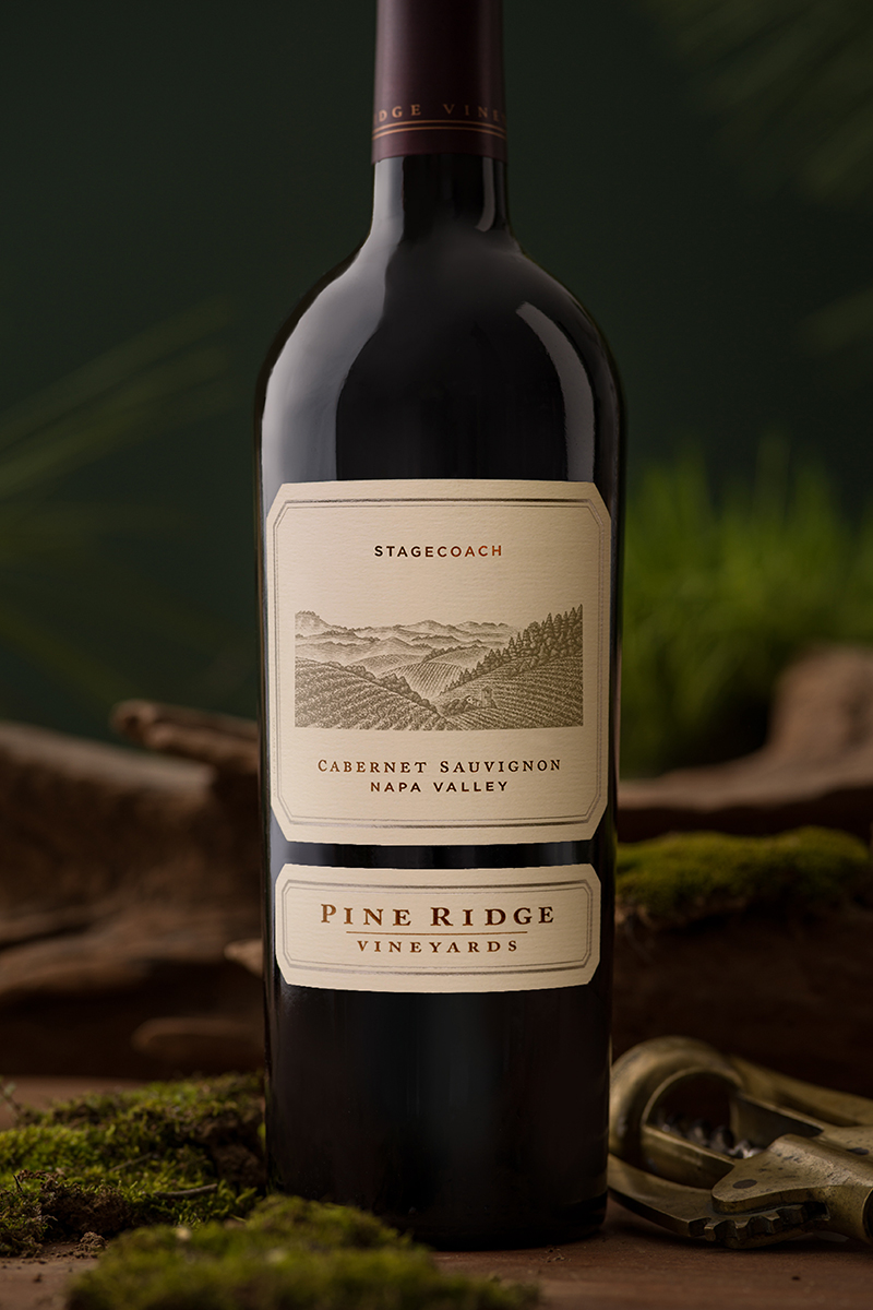 A bottle of Pine Ridge Vineyards Cabernet Sauvignon from Napa Valley. The label reads Stagecoach. The bottle is placed on a surface with moss and wood in the background.