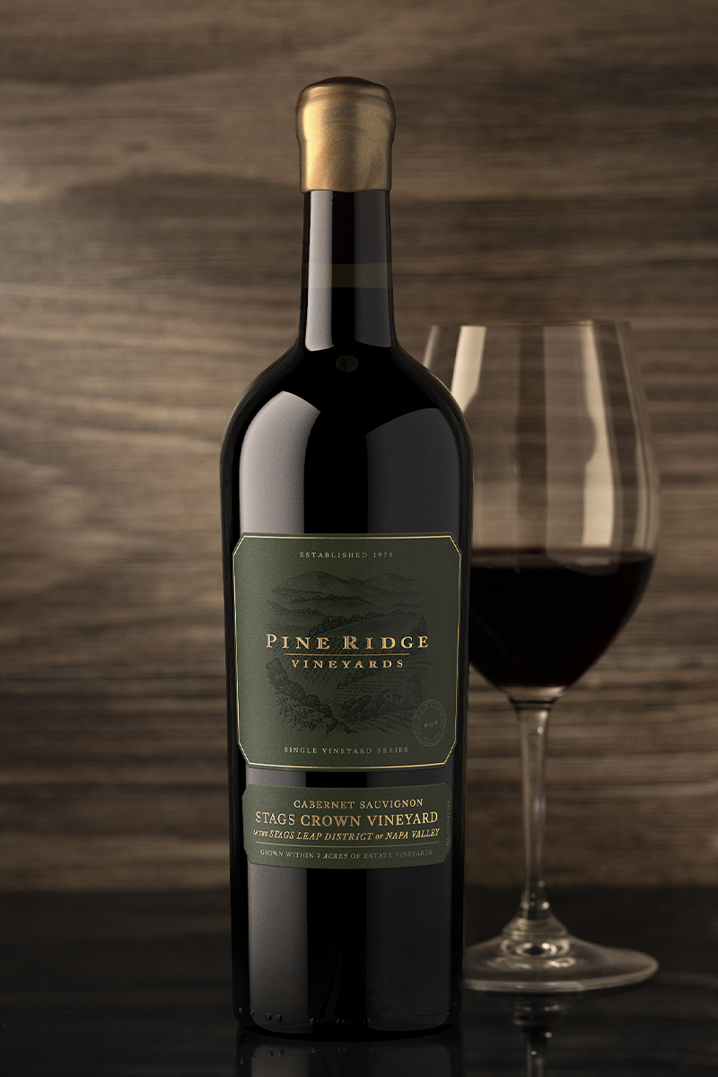 A bottle of Pine Ridge Vineyards Stags Crown Cabernet Sauvignon wine rests on a reflective surface beside a partially filled glass. The label reveals vineyard details, set against a rich wood texture backdrop.