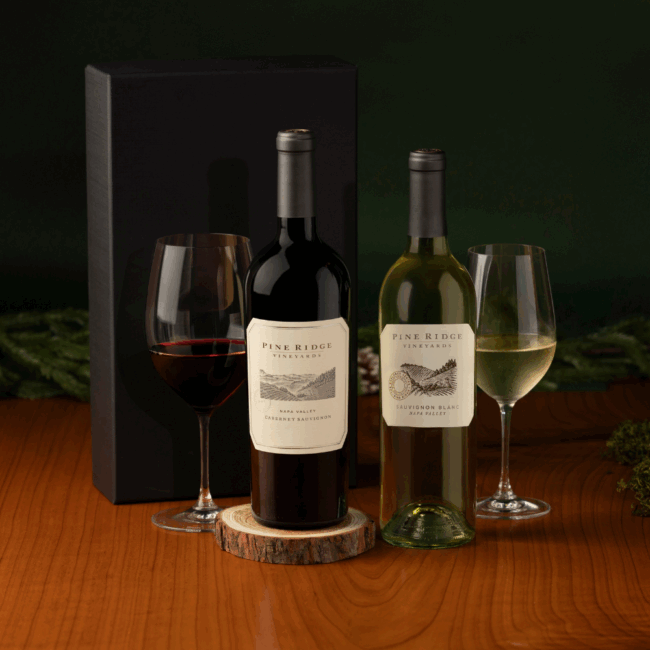 Two bottles of Pine Ridge Vineyards wine, one red and one white, are displayed with matching filled glasses on a wooden surface. A black Napa Valley Favorites 2-Bottle Gift box stands behind the bottles against a dark green background.