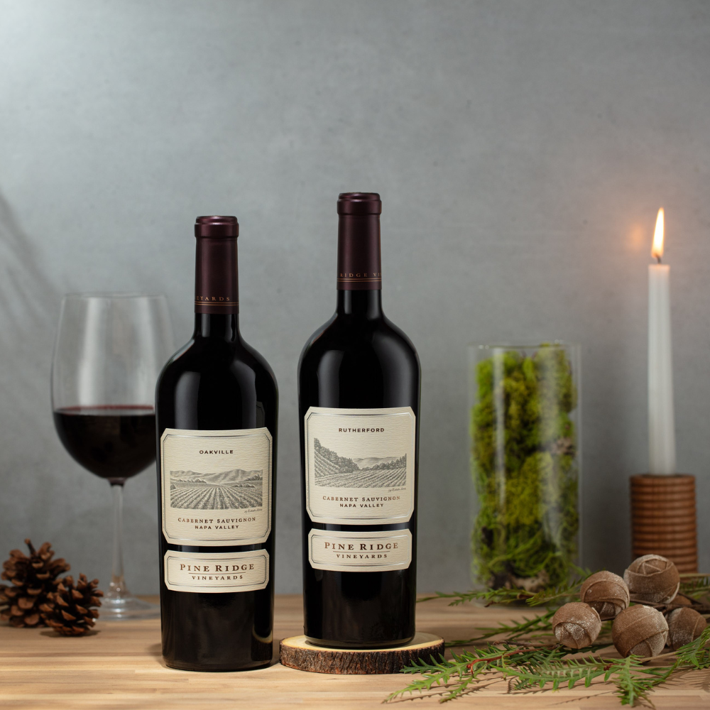 Two bottles from the Napa Valley Appellation 2-Bottle Gift by Pine Ridge Vineyards are displayed on a wooden surface with a wine glass, pinecone, greenery, decorative balls, and a lit candle in the background.
