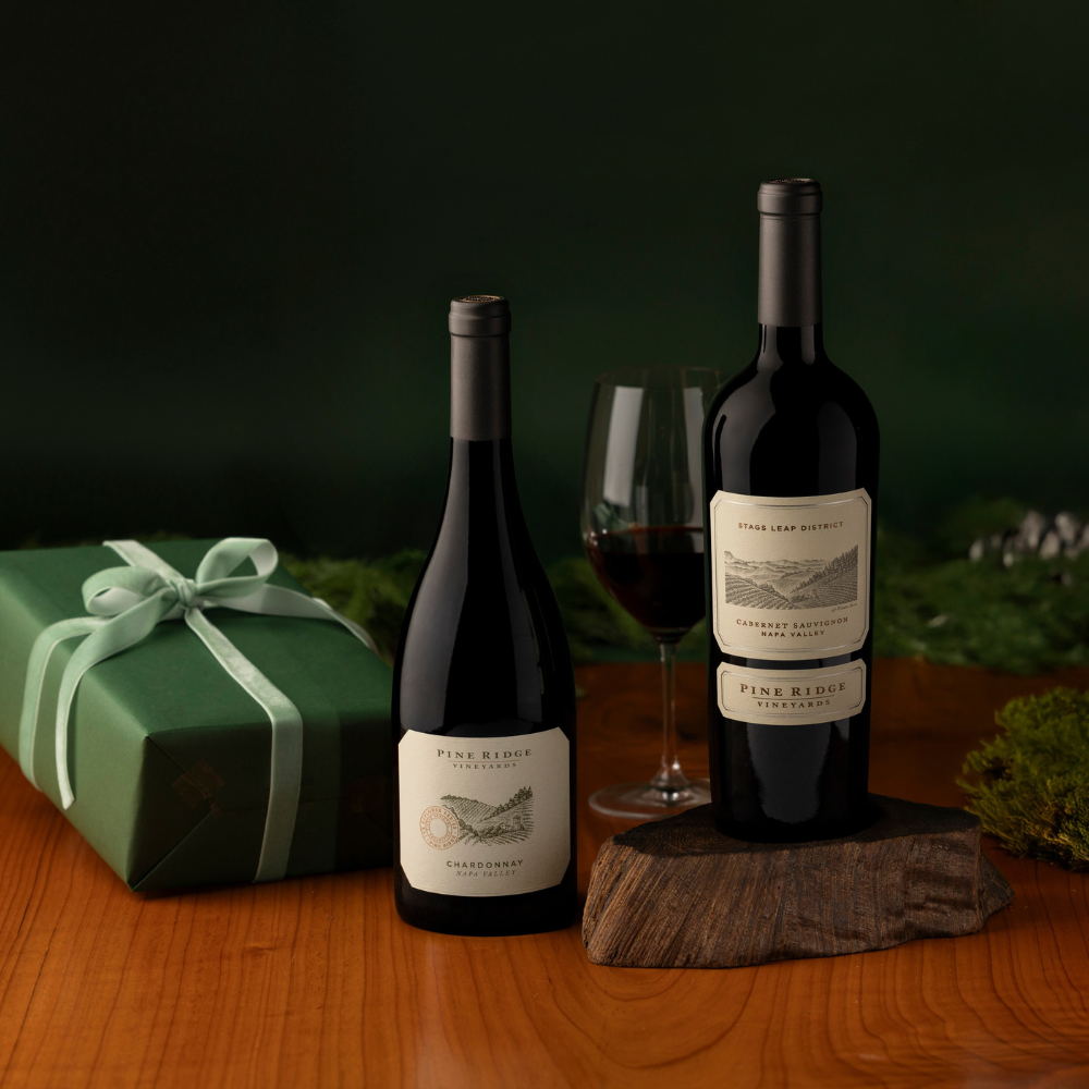 Two bottles of Pine Ridge Vineyards wine—Chardonnay and Cabernet Sauvignon—are displayed on a wooden table with a glass of red wine, a green gift box with ribbon, and greenery, making this the perfect Napa Classics Wine Gift.